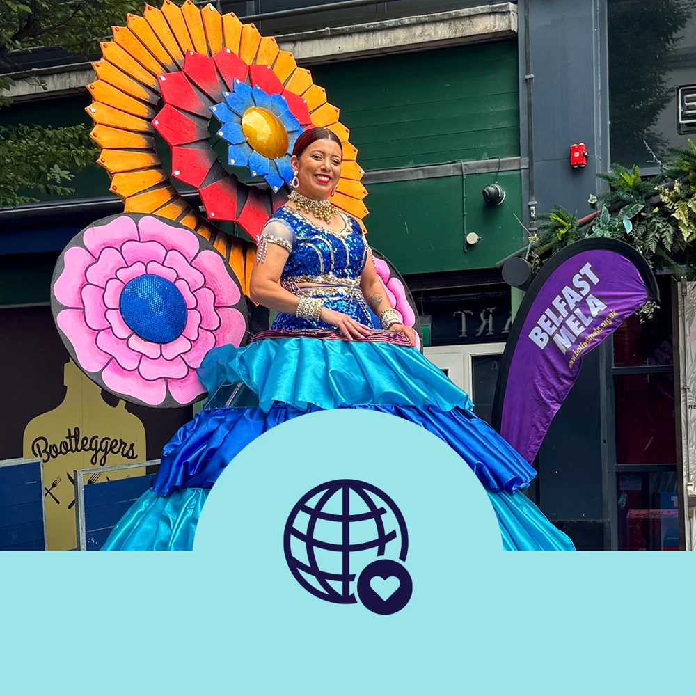A person in a vibrant, elaborate costume featuring large, colorful flowers is participating in the Belfast Mela. They are standing on a float decorated with bright blue and pink elements. A "Belfast Mela" banner is visible in the background.