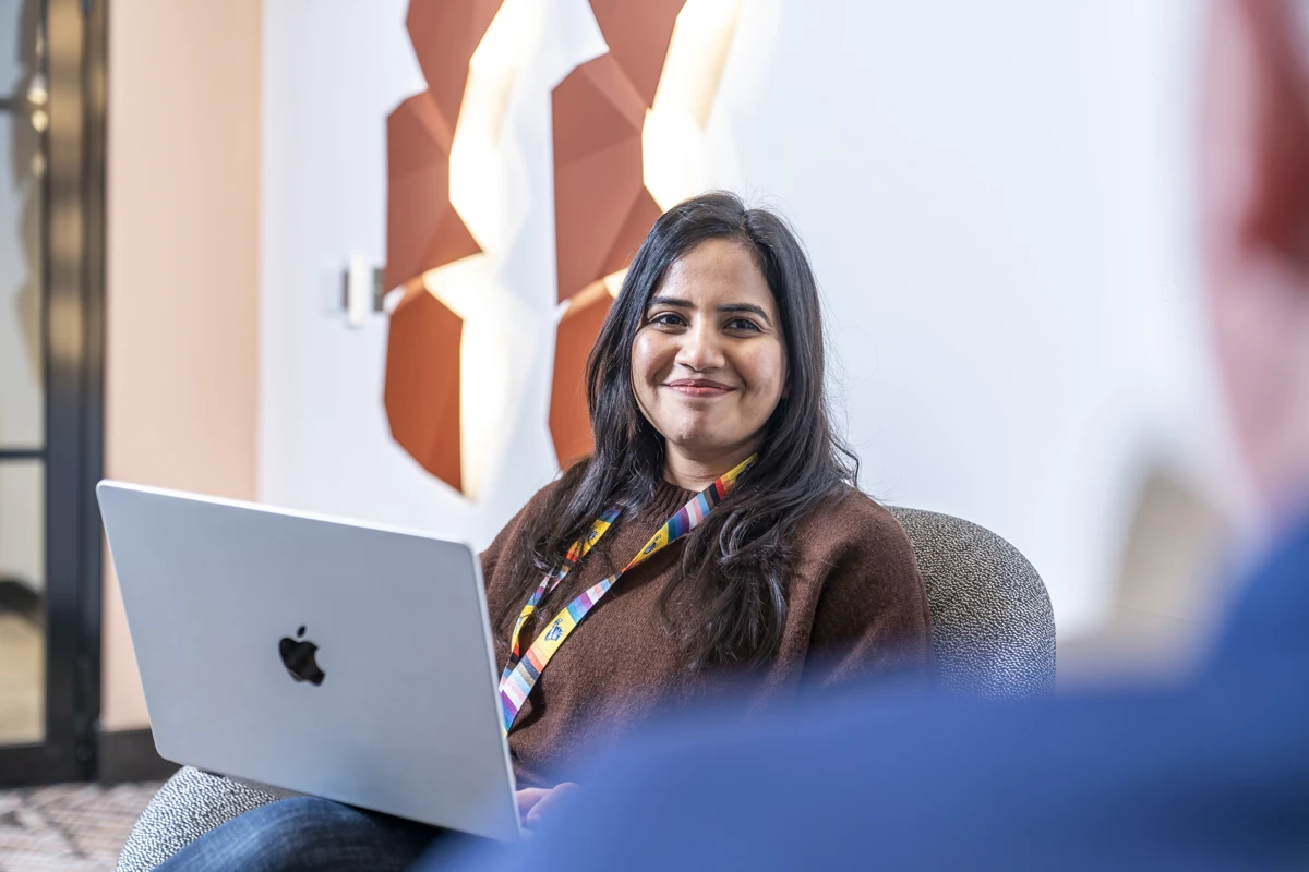 Gouri smiling in an office setting holding her laptop and wearing a Pride lanyard