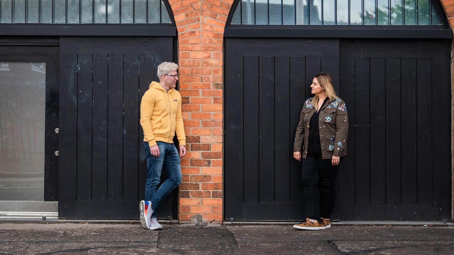 Photo of man and woman talking in front of building with brick wall and black doors