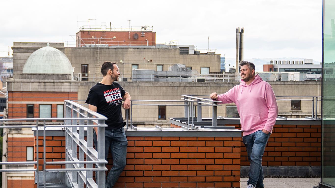 Photo of two men talking on office balcony, with city roofs background