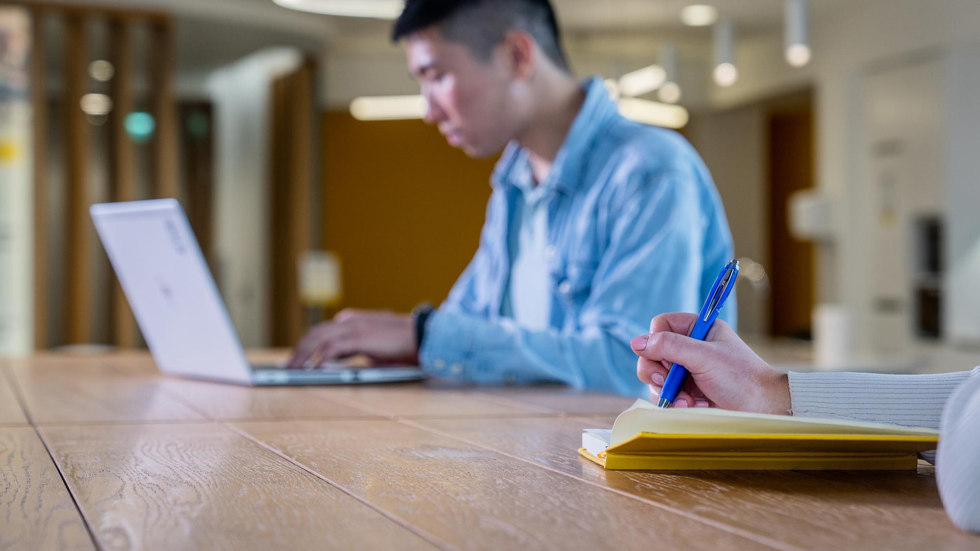 Photo of man at laptop, with woman's hand and notebook in foreground