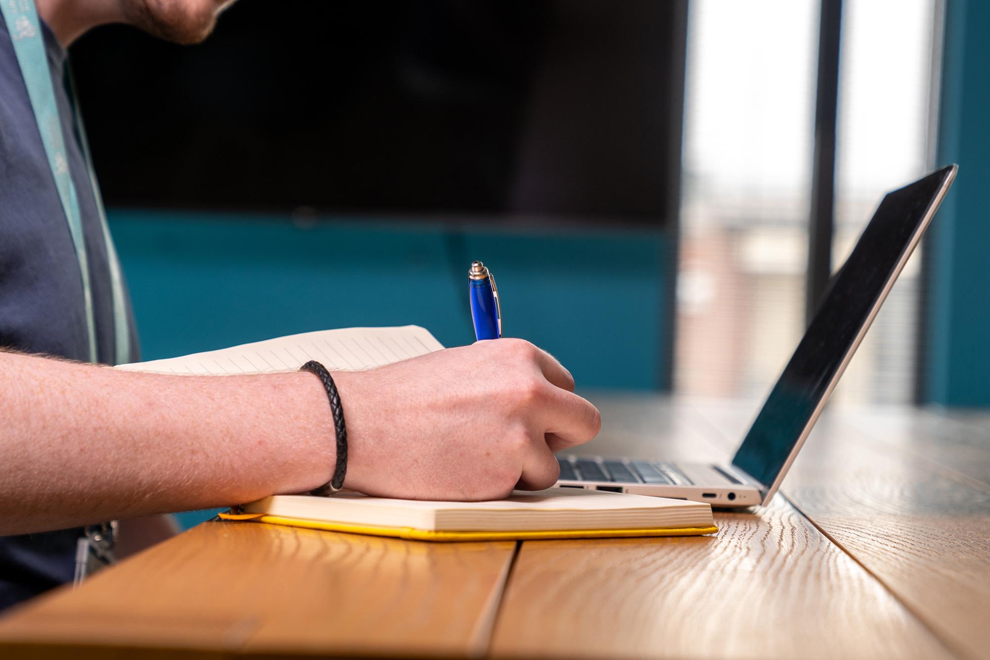 Photo of hand writing on notebook in front of laptop