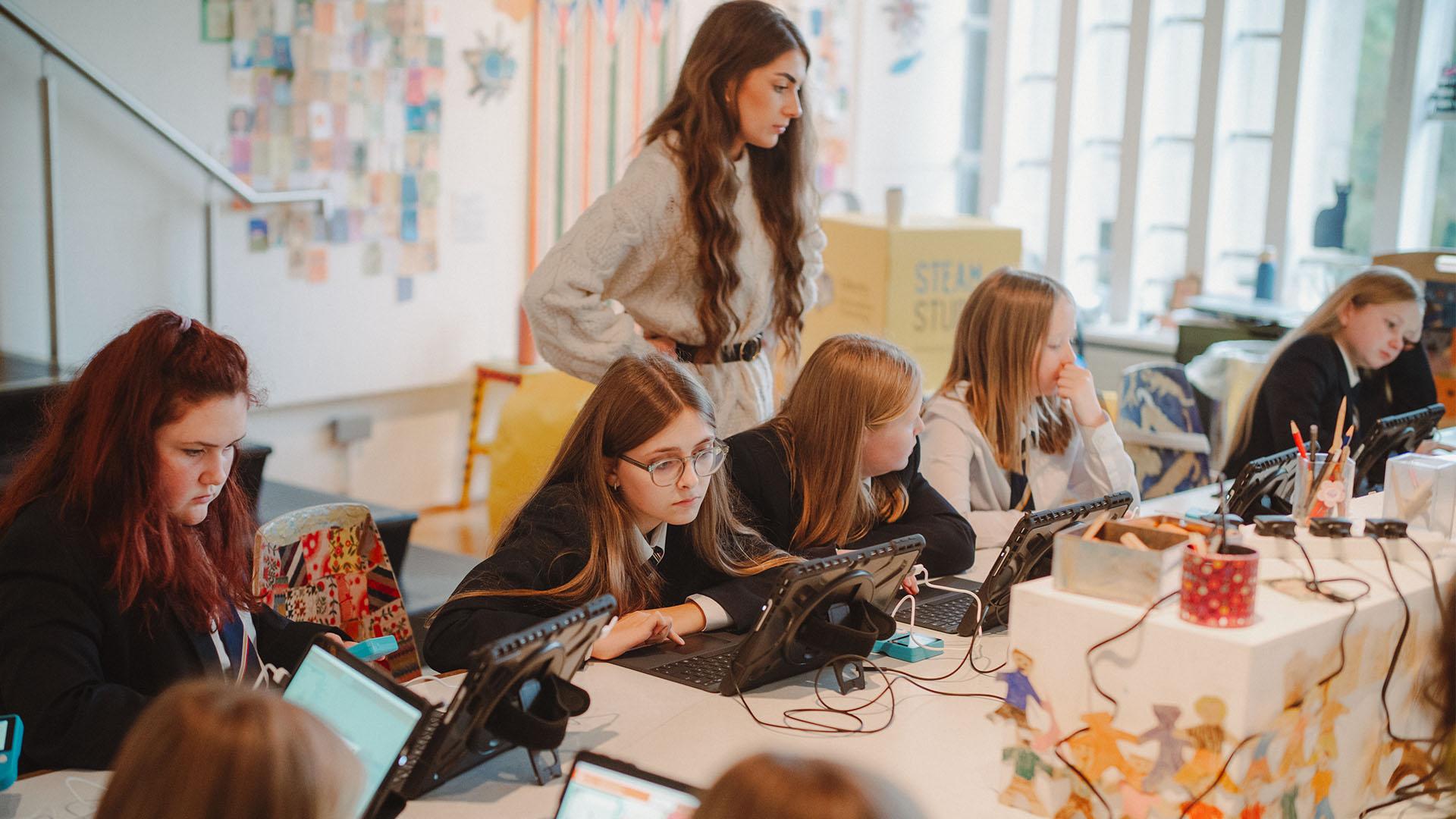 (slide 5 of 9) An adult guiding schoolchildren through a task, with the children sitting at a long desk on laptops.. 