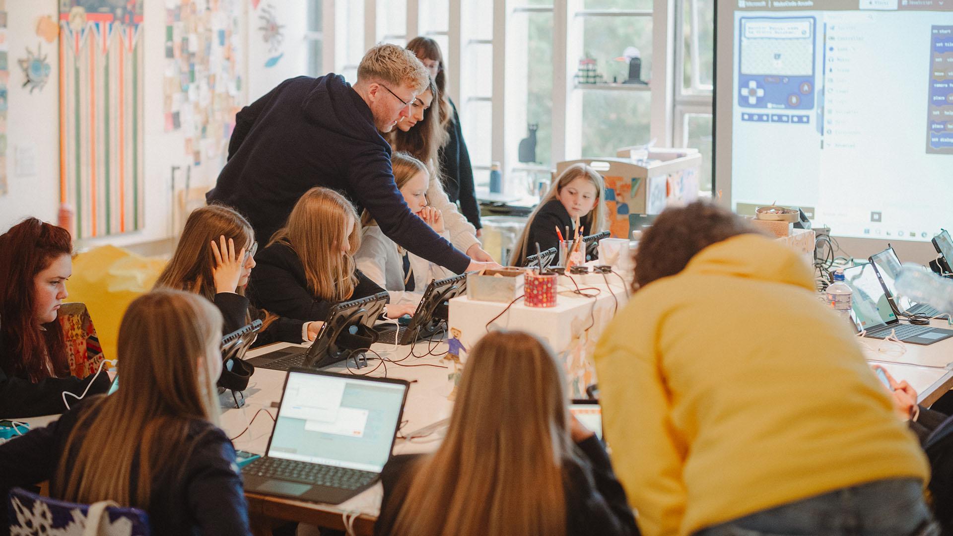 (slide 9 of 9) An adult guiding schoolchildren through a task, with the children sitting at a long desk on laptops.. 