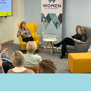 Photo of two women talking in chairs at front of room of people