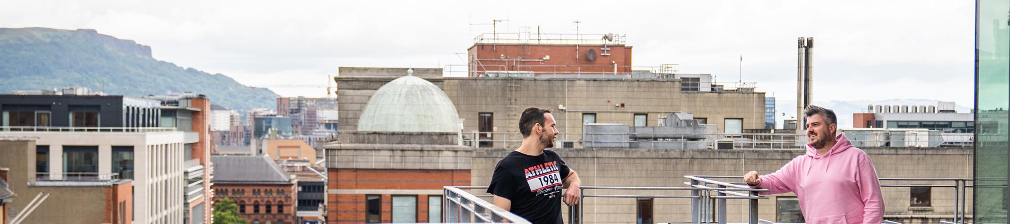 Photo of two men talking on office balcony, with city roofs background